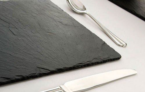 CHARGER GREY SLATE: A grey slate charger plate on a table, accompanied by silverware, ideal for elegant event settings and party occasions. Dimensions: 30cm x 30cm.