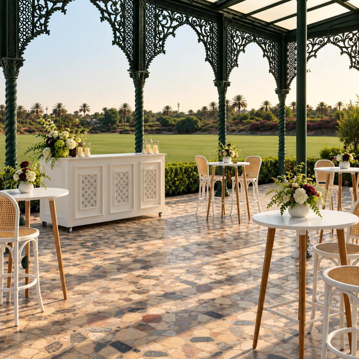 An elegant outdoor patio under a decorative green metal pavilion, featuring white and beige tables, woven bar stools, floral arrangements, and a white serving cabinet. Beyond the patio, a lush green golf course and palm trees are visible under a clear sky.