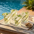 Four glasses of lemonade with lime wedges on a wooden table by a pool.