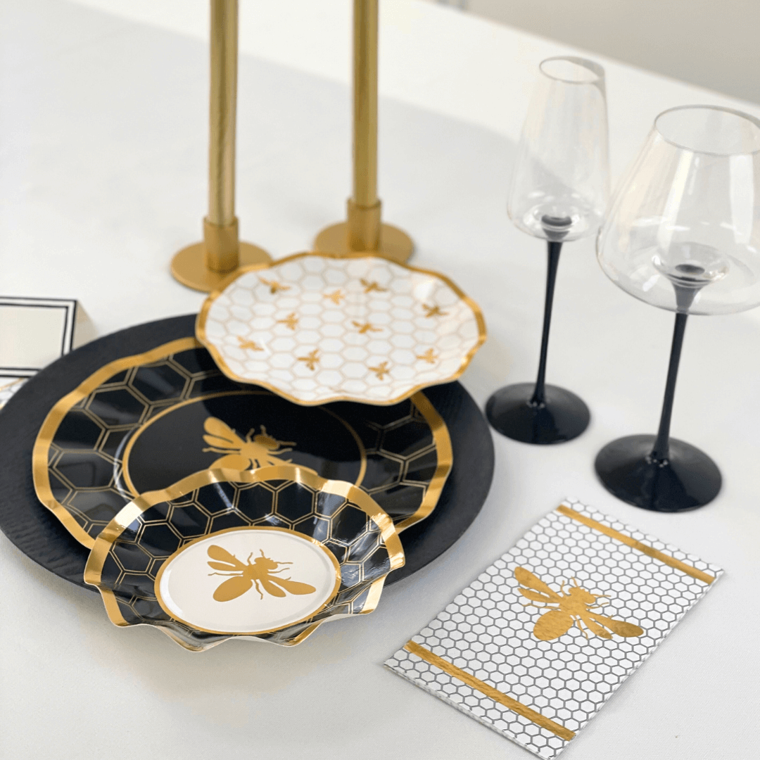 A black and gold plate with a bee design, white and gold card with a bee, and wine glass close-ups. HoneyBee Paper Guest Towel Napkins - 20 per package, ideal for elegant dining setups. From Party Social.