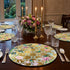 Dining table set with floral placemats, candles, and cutlery in a room with large windows.