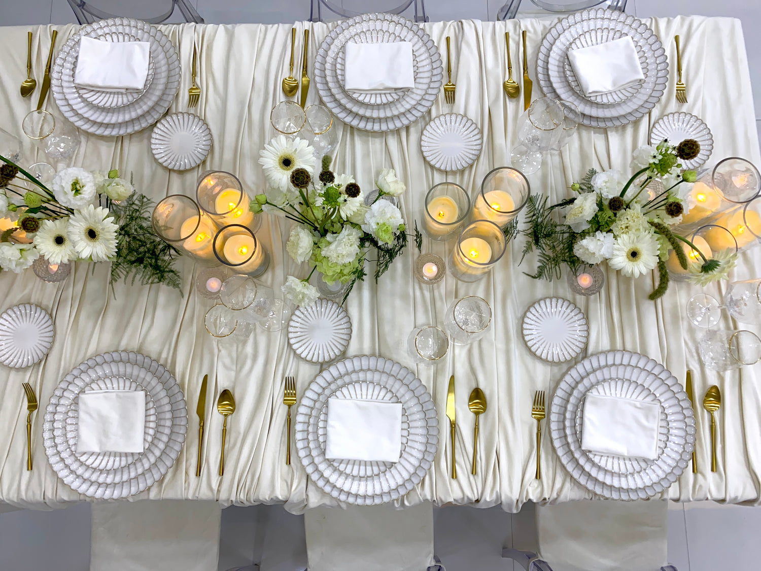 Classic Cream-Dining Setup: Elegantly arranged table for eight, featuring scalloped plates, gold cutlery, glassware, ghost chairs, floral centerpieces, and lit candle vases.