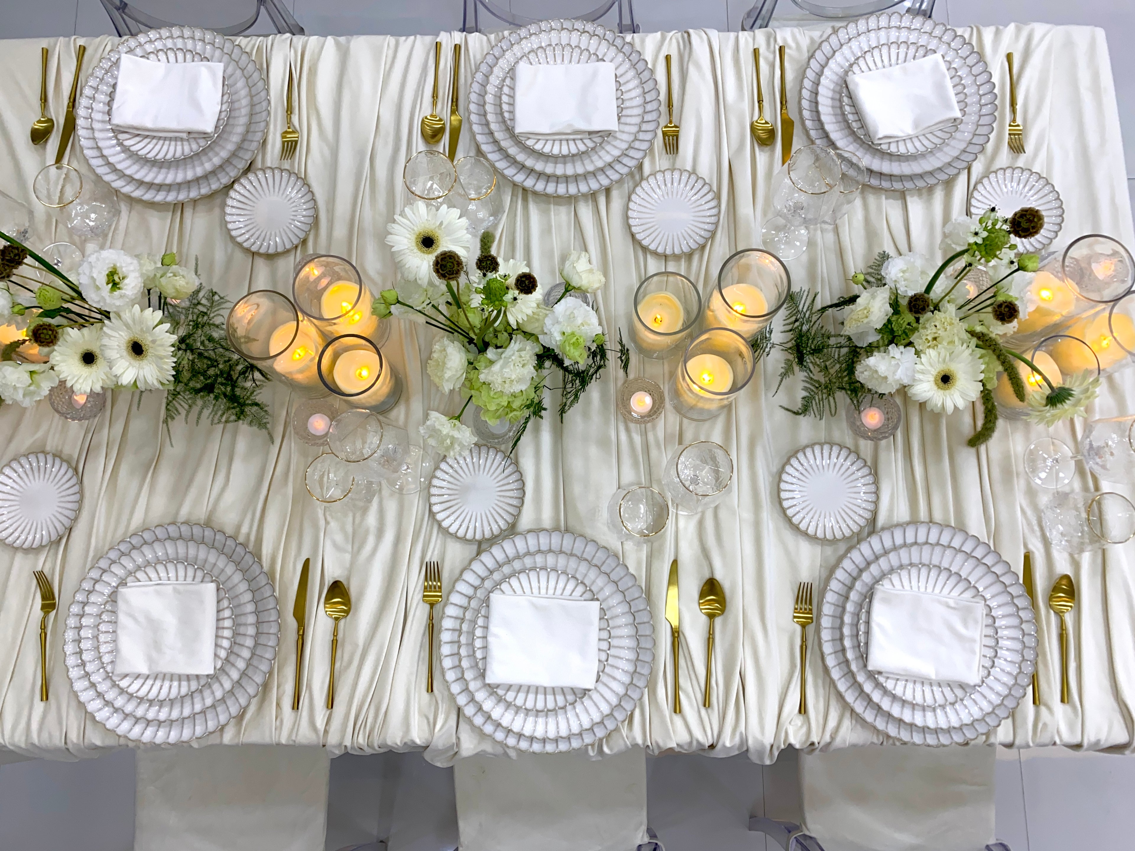 Classic Cream-Dining Setup: Elegantly arranged table for eight, featuring scalloped plates, gold cutlery, glassware, ghost chairs, floral centerpieces, and lit candle vases.