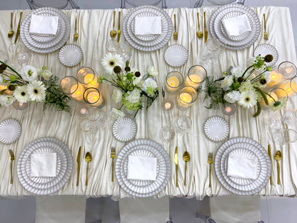 Classic Cream-Dining Setup: Elegantly arranged table for eight, featuring scalloped plates, gold cutlery, glassware, ghost chairs, floral centerpieces, and lit candle vases.