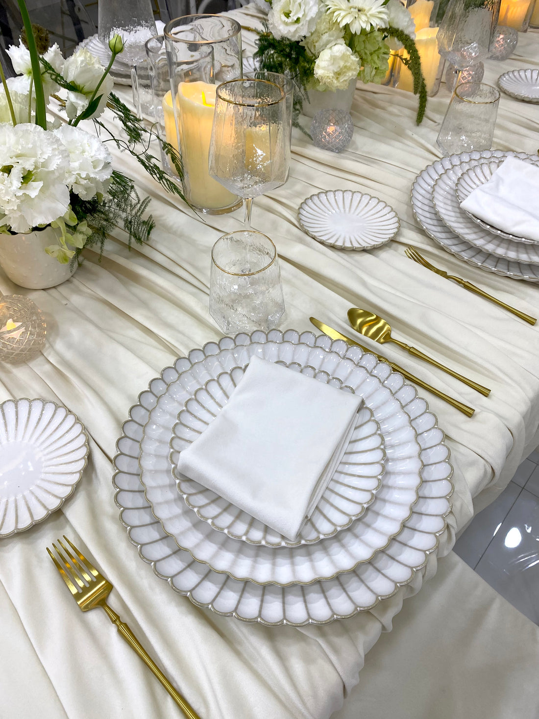 Classic Cream-Dining Setup featuring scalloped-edge plates, gold flatware, crackle-textured glasses, and flower arrangements on a draped tablecloth, perfect for elegant occasions.