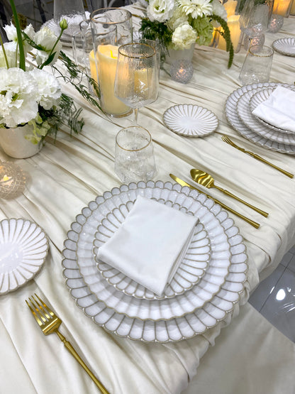 Classic Cream-Dining Setup featuring scalloped-edge plates, gold flatware, crackle-textured glasses, and flower arrangements on a draped tablecloth, perfect for elegant occasions.