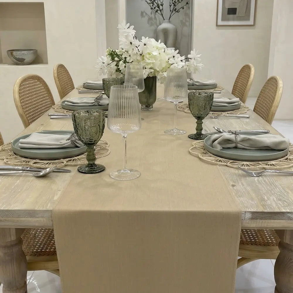 LINEN BLEND TABLE RUNNERS adorn the light-wood table, enhancing a six-setting arrangement with woven placemats, muted green plates, and vintage-style goblets for elegant occasions.
