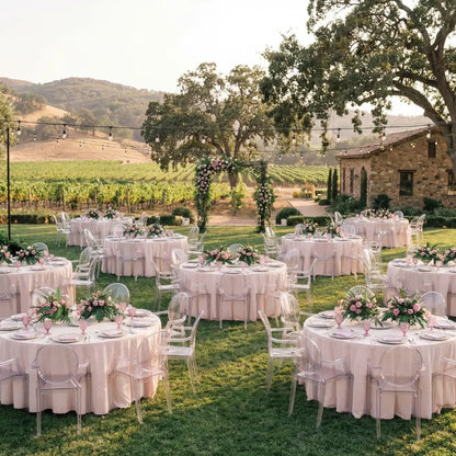 GHOST CHAIR WITH ARMS (LOUIS) elegantly complements a vineyard wedding setup, encircling tables with pink decor under string lights, highlighting modern luxury in a rustic setting.