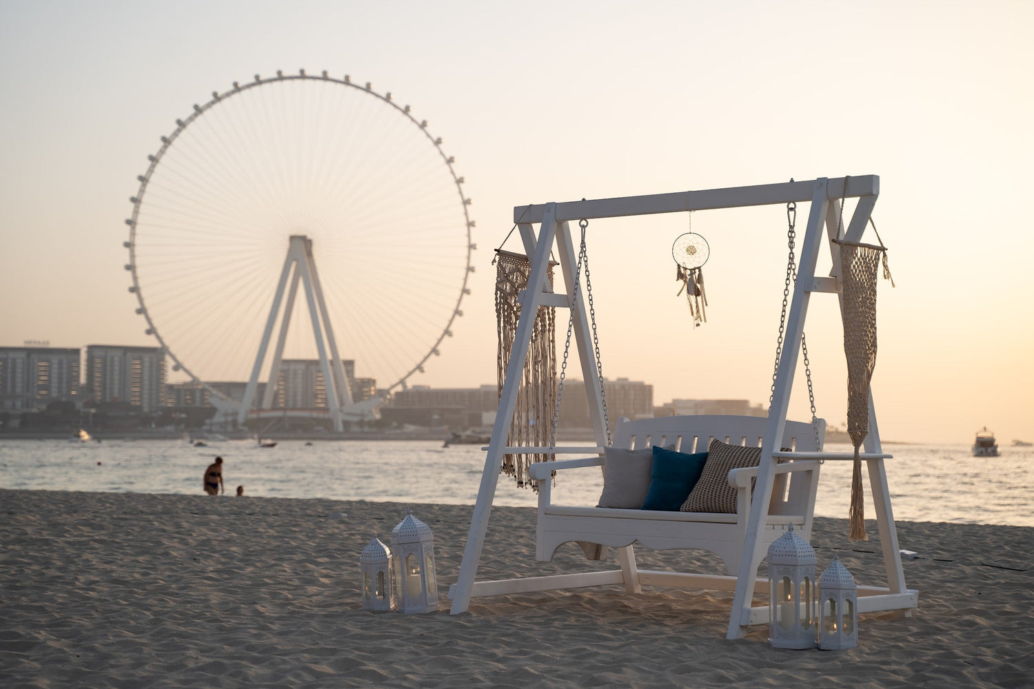 White Wooden Swing on a beach, perfect for event decor or photography, with a Ferris wheel in the background. Ideal for rental or purchase.