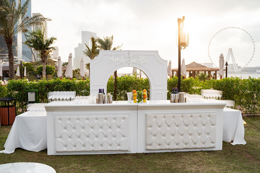 White leather bar unit with a sleek design, ideal for indoor or outdoor party setups. Featured in a garden setting, showcasing its elegant versatility. Dimensions: 185cm x 65cm x 107cm.