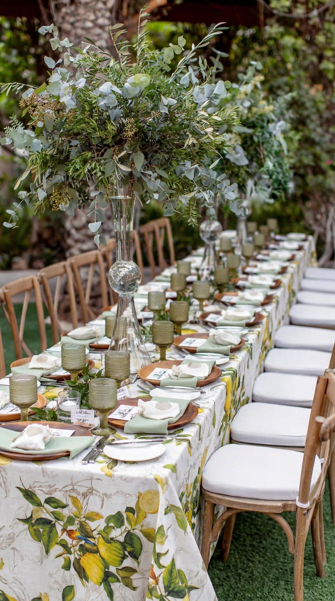 Limoncello Citrus Pure Linen Tablecloth drapes a banquet table, featuring lemon and leaf motifs, complemented by wooden chargers, sage napkins, and vintage green goblets for elegant outdoor dining.