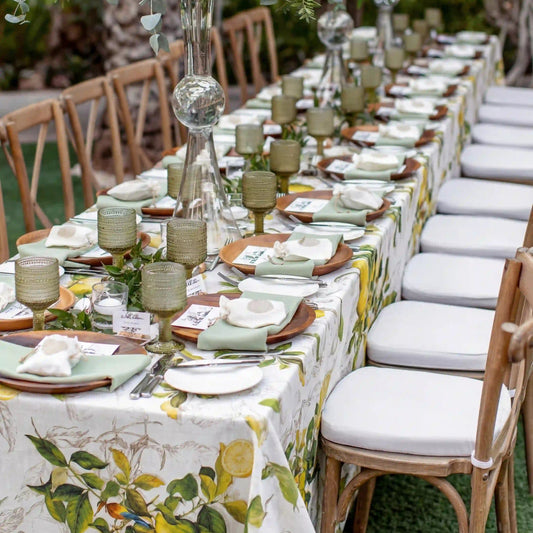 Limoncello Citrus Pure Linen Tablecloth drapes a banquet table, featuring lemon and leaf motifs, complemented by wooden chargers, sage napkins, and vintage green goblets for elegant outdoor dining.