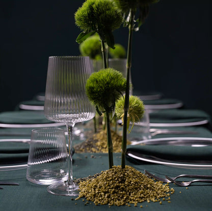 Clear Lined Glassware Collection on a green tablecloth, featuring ribbed wine and water glasses, complementing a minimalist centerpiece with sculptural, chartreuse-green flowers.