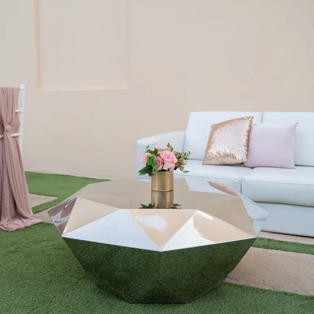 Mirrored silver coffee table with a reflective finish, part of a stylish lounge setup, featuring a vase of pink roses, ideal for event settings.