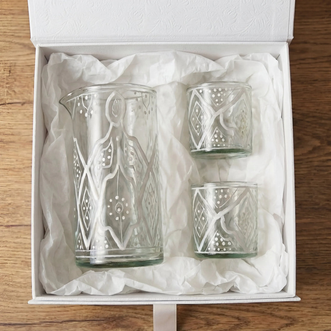 Marrakesh Glassware Gift Box with a clear glass carafe and two tumblers, featuring white geometric patterns, elegantly presented in a white gift box on wood.