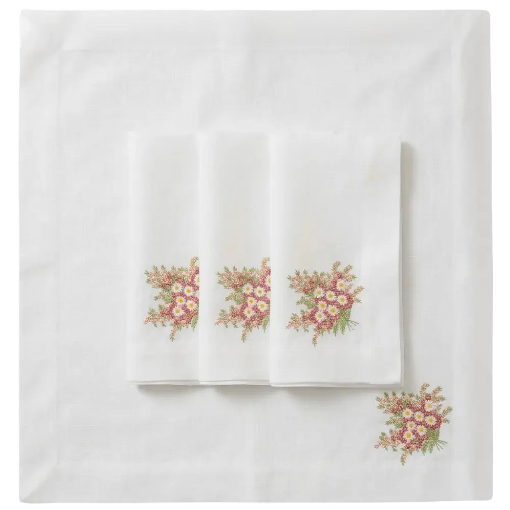White tablecloth with three folded napkins, each featuring embroidered pink and white daisies with greenery, highlighting the Pink Daisy Pure Linen Dinner Napkin - Set of 4.
