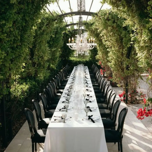 TABLE 6' RECTANGULAR BANQUET set elegantly outdoors, draped in white, surrounded by black chairs under a pergola with vines and a chandelier.