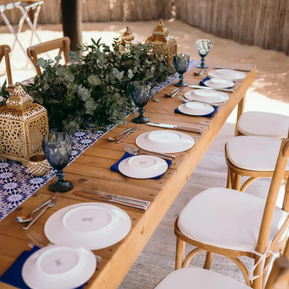 RECYCLED WOODEN DINING TABLE set for an outdoor event, featuring a lush foliage garland, brass lanterns, and elegant tableware, surrounded by simple wooden chairs.