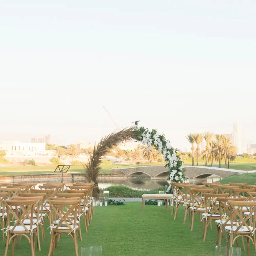 Round Gold Arch set up outdoors by a pond, adorned with white flowers and palm fronds, enhancing a wedding ceremony setup with wooden chairs.