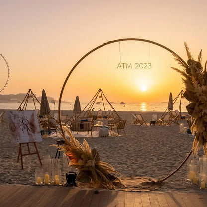 Round Gold Arch adorned with dried grasses and florals, set on a beach with lounge chairs and pillar candles, ideal for elegant event backdrops.