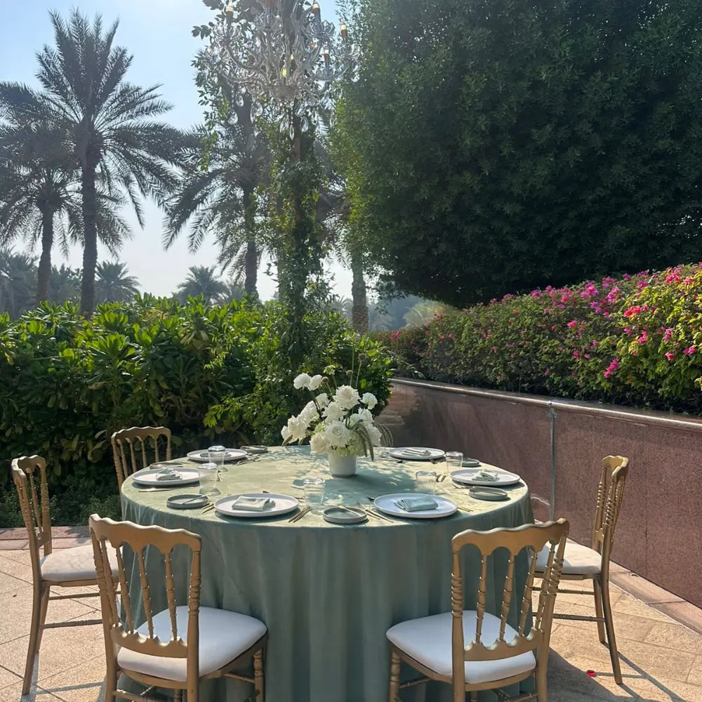 Round Banquet Table set for six with white plates, gold-tone cutlery, and floral centerpiece, surrounded by wooden chairs with cushions in an elegant garden setting.
