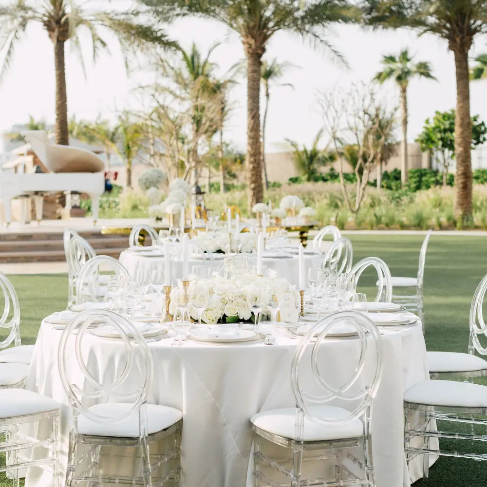 Elegant wedding setup featuring a ROUND TABLECLOTH SATIN on a round table, adorned with white florals and candles, surrounded by ghost chairs on a lawn.
