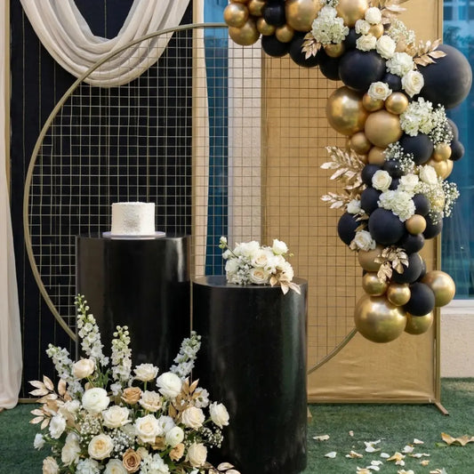 Round White Podiums displaying a small white cake and floral bouquet, set against an elegant backdrop with black and gold balloons, perfect for upscale events.