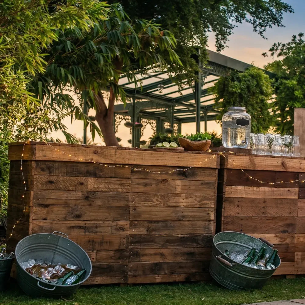 Rustic Bar Unit in garden setting with wooden design, fairy lights, beverage dispenser, herbs, limes, and glasses, ideal for outdoor events.