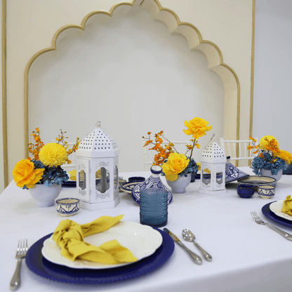 Moroccan Mirage- Dining Setup featuring a table with white Moroccan lanterns, blue and white plates, gold cutlery, and floral centerpieces for eight guests.