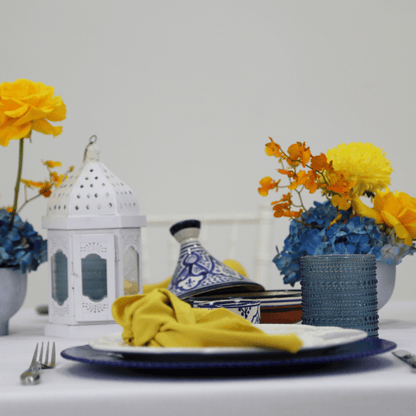 Moroccan Mirage- Dining Setup featuring a table with blue and white plates, gold cutlery, white lanterns, floral centerpieces, and glassware for eight guests.