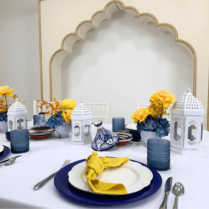 Moroccan Mirage- Dining Setup featuring a table set for eight with blue and white pottery, gold cutlery, floral centerpieces, and white Moroccan lanterns.