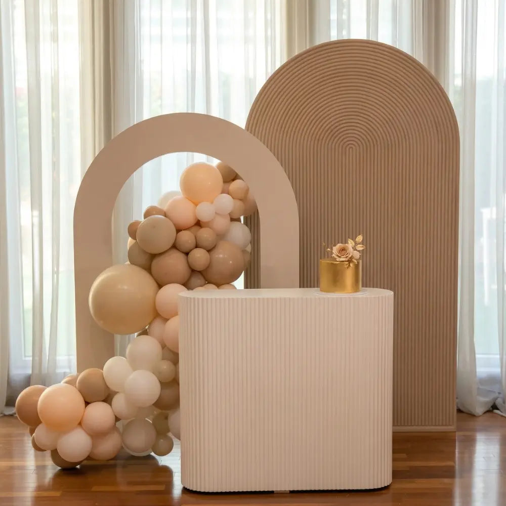 Rippled White Counter Podium with vertical ribbing complements event decor, featuring a gold cake and balloon arch, ideal for party setups by Party Social.