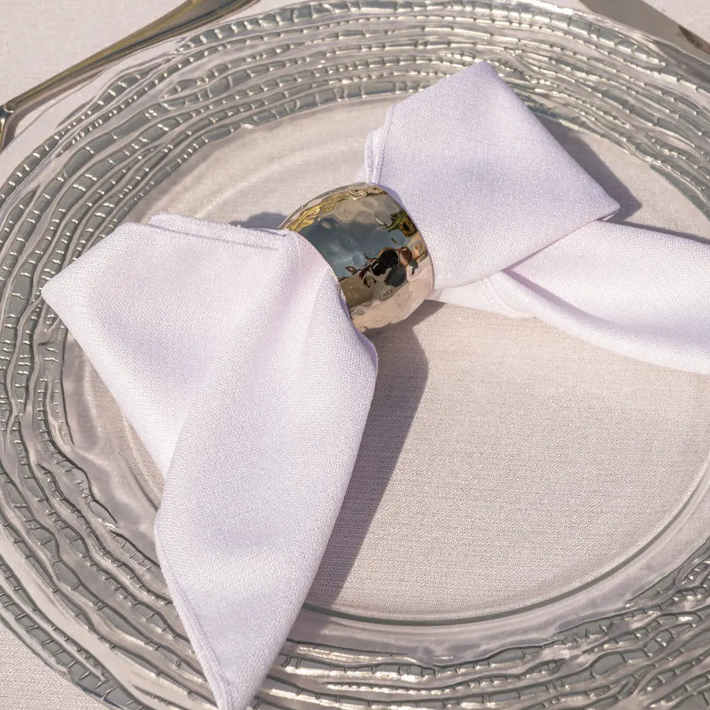 NAPKIN RING - HAMMERED DOME: A hammered gold napkin ring in a bow shape on a white cloth napkin, displayed on a clear glass charger plate.