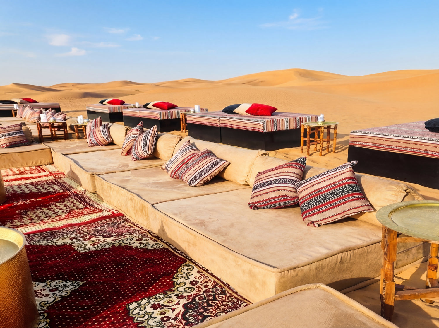 LOUNGE LOW SEATING on sand dunes with cushioned platforms, patterned pillows, and wooden side tables, creating an inviting, intimate gathering space.