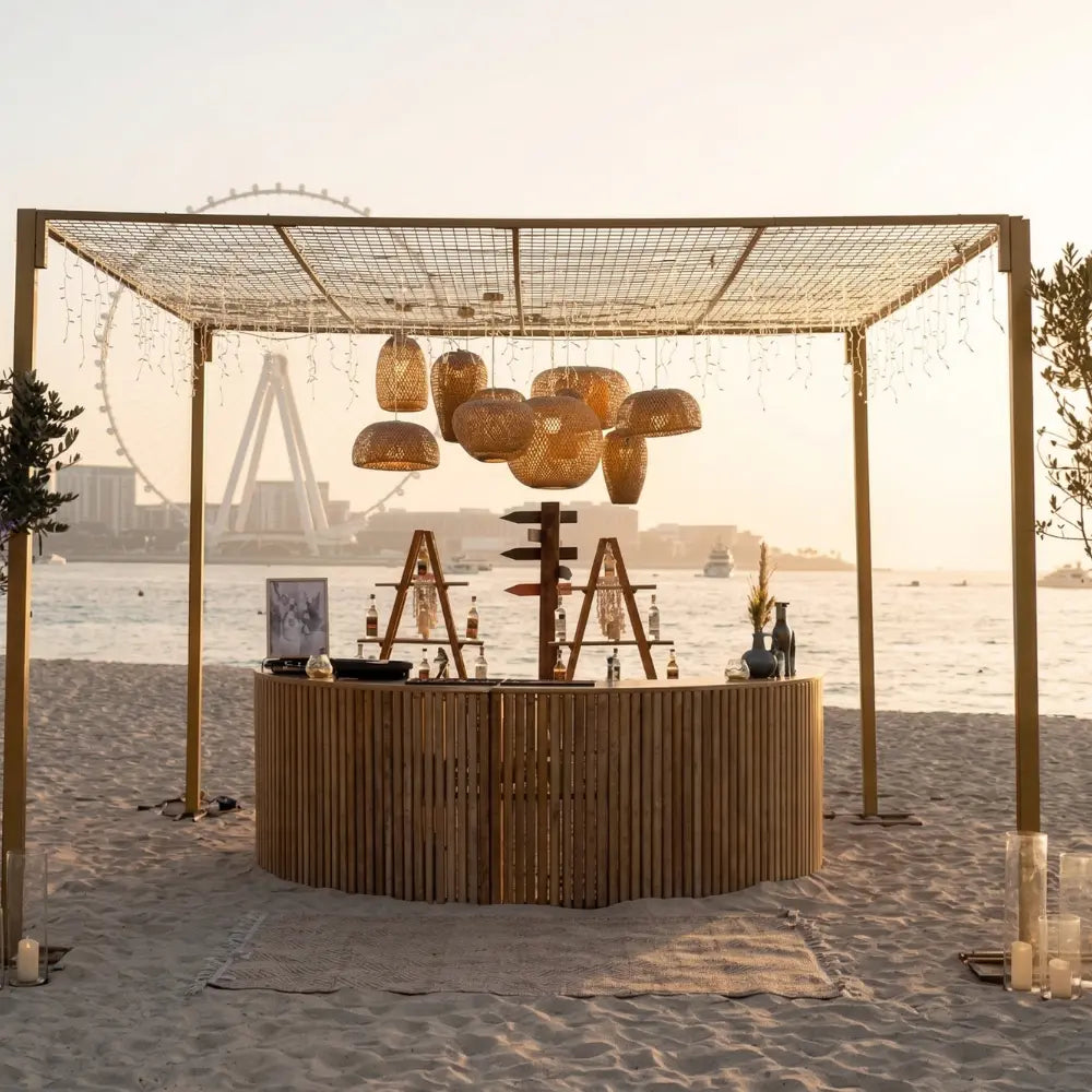 SEMI WOODEN BAR UNIT at a beach setting under a pergola with string lights, featuring bamboo slats and shelves, ideal for garden or home events.