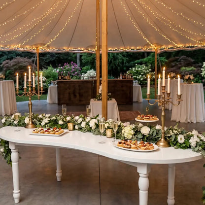 White Crescent Dining Table elegantly set with greenery, brass candelabras, hors d’oeuvres, and sparkling wine, under a canopy at an event reception.