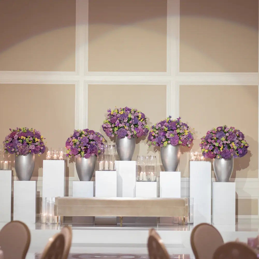 Square White Podiums display floral arrangements on staggered pedestals, enhancing event aesthetics with metallic vases and blooms, ideal for weddings and formal gatherings.