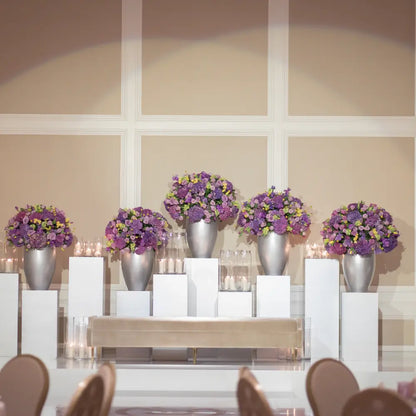 Square White Podiums display floral arrangements on staggered pedestals, enhancing event aesthetics with metallic vases and blooms, ideal for weddings and formal gatherings.