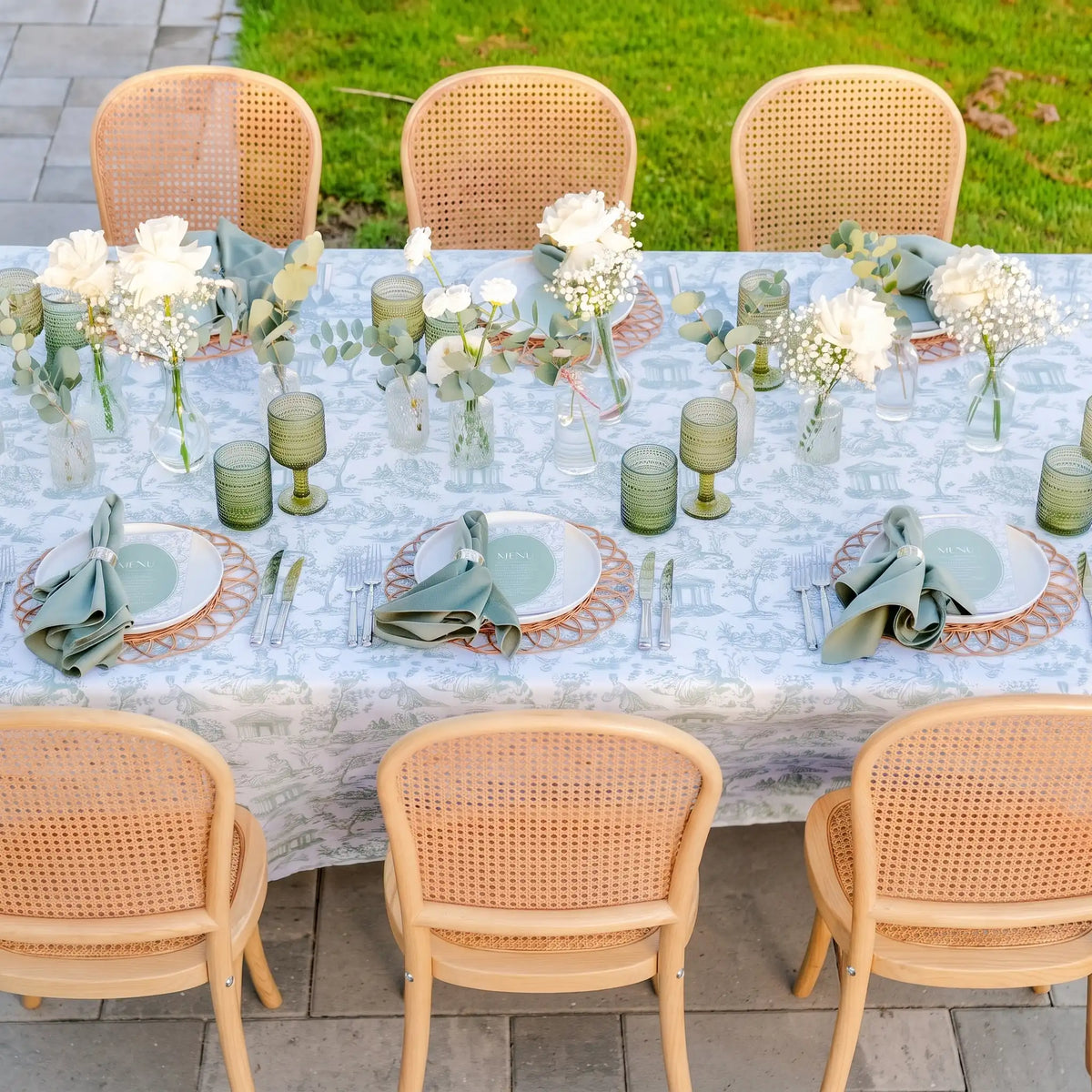 An outdoor dining table set with a light blue and white toile-print tablecloth, white plates, sage green napkins, white flowers, and green glass candlesticks, surrounded by woven cane chairs.