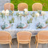An outdoor dining table set with a light blue and white toile-print tablecloth, white plates, sage green napkins, white flowers, and green glass candlesticks, surrounded by woven cane chairs.