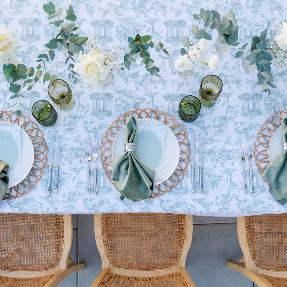 A styled dining table setup with a light blue and white toile patterned tablecloth, white plates, green napkins, woven gold placemats, green glassware, and floral centerpieces, viewed from above with three woven chairs visible below.
