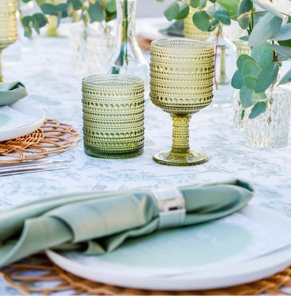 A styled dining table setting featuring textured, ribbed glassware in sage green and olive tones, paired with white plates, folded sage green napkins, and woven placemats.