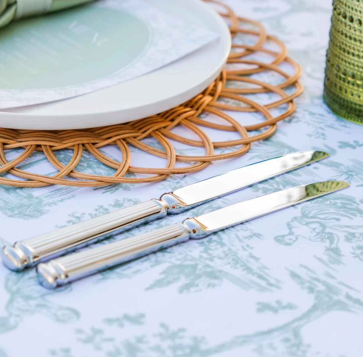 Two silver butter knives with ribbed handles resting on a light blue and white toile patterned tablecloth, styled with a woven placemat, white plates, and a green textured candle.