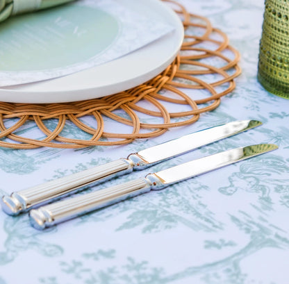 Two silver butter knives with ribbed handles resting on a light blue and white toile patterned tablecloth, styled with a woven placemat, white plates, and a green textured candle.