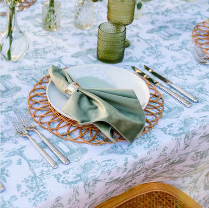 A styled table setting featuring a white tablecloth with a light blue toile de Jouy pattern, a white charger plate, a woven round gold charger, and a sage green satin napkin tied with a gold ring.