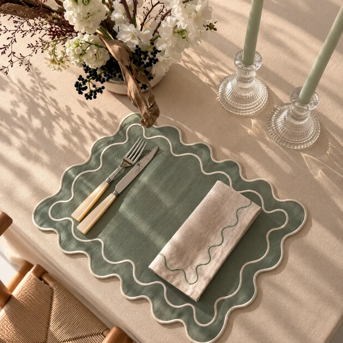 A scalloped sage green placemat with a white border, styled on a beige tablecloth with a folded cream napkin, silver cutlery with light wood handles, and a floral centerpiece under soft window shadows.