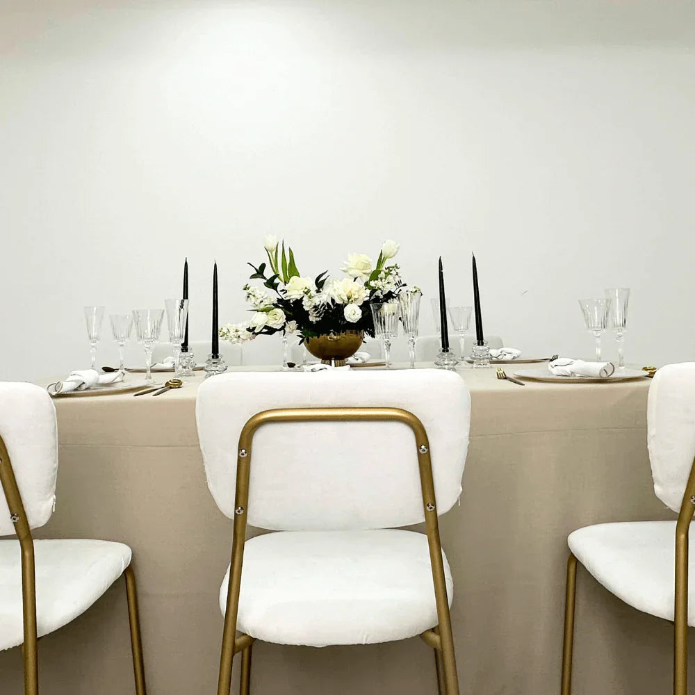 Dining table set with white tablecloth, floral centerpiece, and gold chairs.