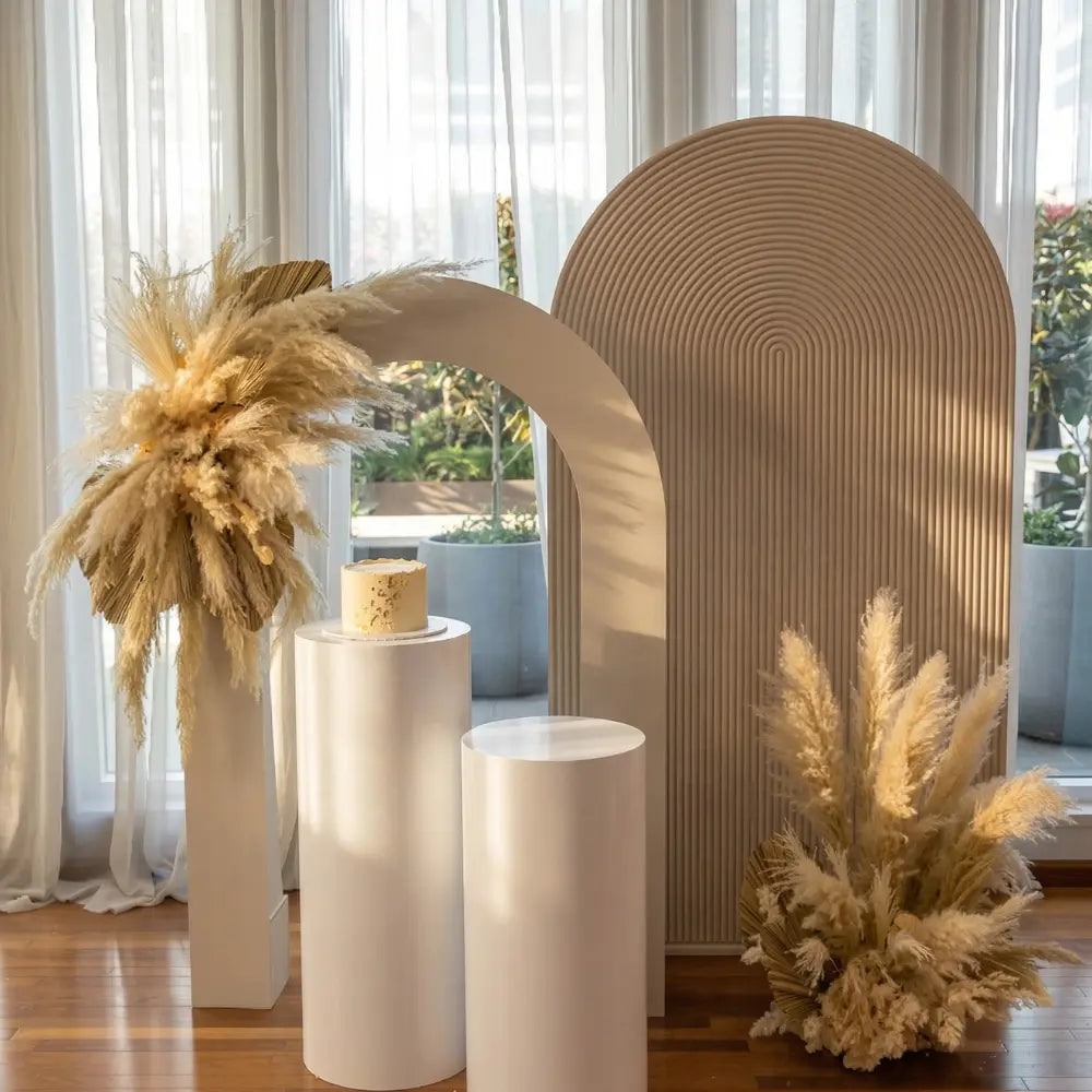 Sand Molded Backdrop with freestanding arches and pampas grass, perfect for elegant event settings, enhanced by cylindrical pedestals for display.