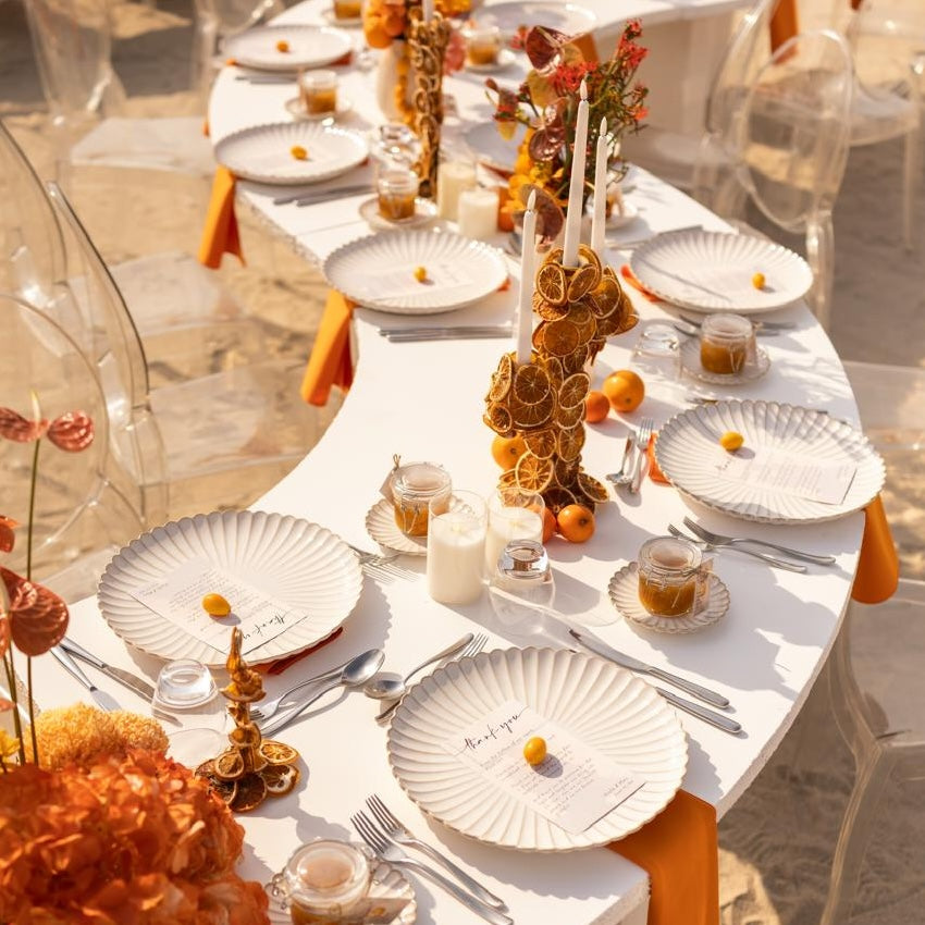 Scalloped Like Plates elegantly arranged with citrus accents on a beachside table, featuring fan-fluted design, silver cutlery, and orange-hued decor for a festive atmosphere.