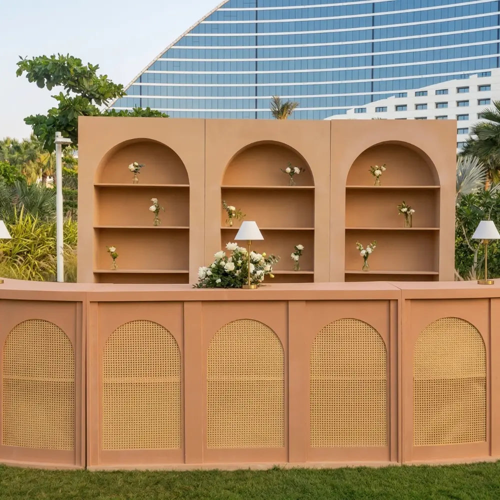 Wooden Rattan Bar Unit with woven cane arch panels and matching arch-topped shelves, adorned with white floral arrangements and table lamps, perfect for elegant event settings.